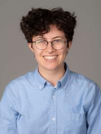 Headshot of St. Clare, a white butch with curly brown hair, glasses, and a blue button-down shirt.