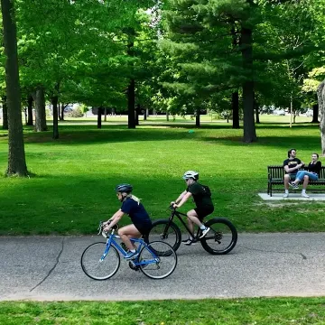 Two students bike on a bike trail through a green park.