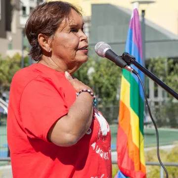 A woman wearing a red shirt, with her hand on her heart, stands aside a rainbow flag.