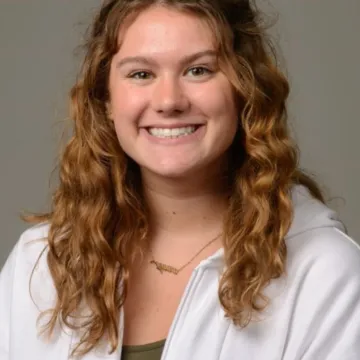 A female presenting person with shoulder-length curly hair. She is wearing an open white shirt over a black top. She is smiling.