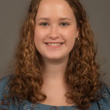 A female presenting person with shoulder-length curly hair. She is wearing gold hoop earrings and a blue floral top. She is smiling.