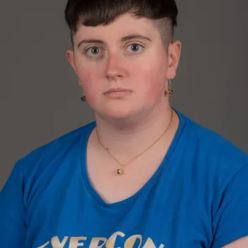 A nonbinary person with short brown hair. They are wearing gold dice jewelry and a blue T-shirt that says EVERCON 24.