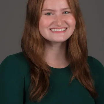 A female presenting person with shoulder length wavy red hair. She is wearing a green long sleeve top and is smiling.