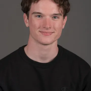 A white male presenting person with short brown hair. He is wearing a black top and is smiling.