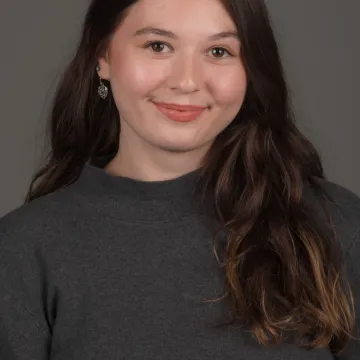 A white female presenting person with long brown hair. She is wearing heart earrings and a gray turtleneck.