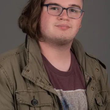 A nonbinary person with chin-length brown hair that lays to one side. They are wearing glasses, a green jacket and a maroon shirt with a wave print.