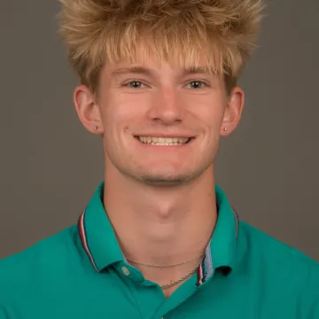 A white male presenting person with blonde spicky hair. He is wearing a green polo and is smiling.