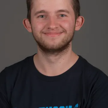 A white male presenting person with short brown hair and a small bear. He is wearing a blue shirt and smiling.