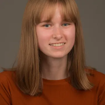 A white female presenting person with shoulder length blonde hair. She is wearing a red top and smiling.