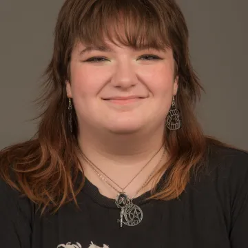 A nonbinary person with brown shoulder length hair and bangs. They are wearing an assortment of silver jewelry and a band T-shirt. They are smiling. 