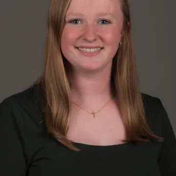 A female presenting person with shoulder length blonde hair. She is wearing gold jewelry and a black top. She is smiling.