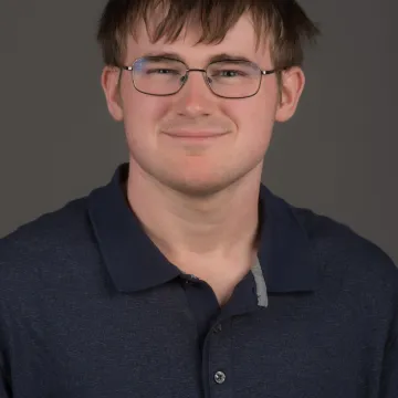 A male presenting person with short brown hair. He is wearing glasses and a polo. He is smiling.