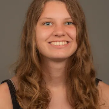 A white female presenting person with long wavy brown hair. She is wearing a black sleeveless shirt and is smiling.