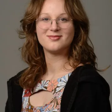 A female presenting person with brown wavy hair. She is wearing glasses, a floral print top, and a black jacket. She is smiling.