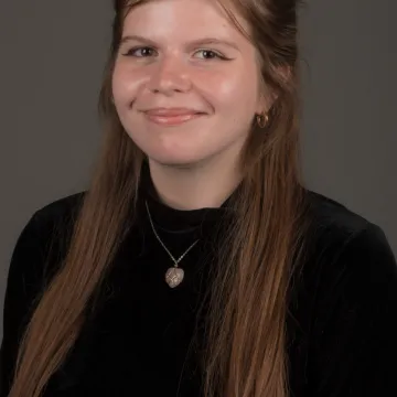 A female presenting person with long blonde hair. She is wearing gold jewelry and a black top. She is smiling.