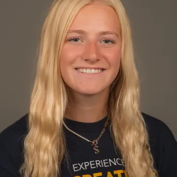 A white female presenting student with long blonde hair. She is wearing a necklace and UWEC shirt. She is smiling.