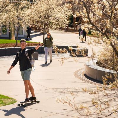 male student on a long board crossing campus 