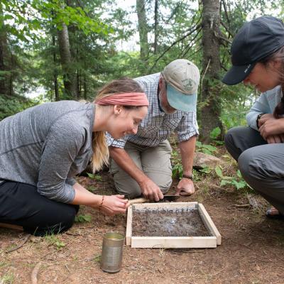 Group of researchers examining a soil sample in a forested area, using a wooden frame and metal can, while kneeling on the ground and focusing on their outdoor fieldwork.