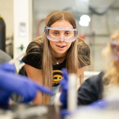 A student wearing safety goggles intently observing a laboratory experiment, with other students working alongside, focusing on the scientific process.