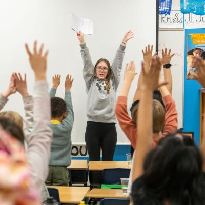 A teacher leads a classroom activity with energetic elementary school children enthusiastically raising their hands.