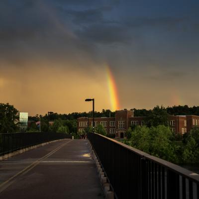 A rainbow emerges from the clouds and appears to end on Schofield Hall as seen from the footbridge.