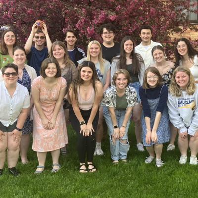  The image shows a group of fifteen young adults posing outdoors on a grassy area, with a flowering tree and brick building in the background. They are smiling and dressed casually. One person in the back is holding a small orange object above their head.