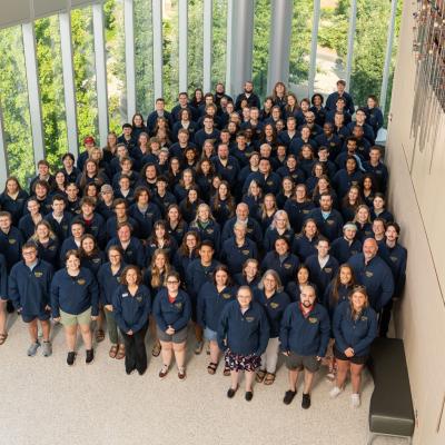 A large group of people in matching navy blue jackets poses for a group photo in a bright indoor space with tall windows and greenery outside.