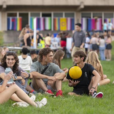 Students sit and chat on the grass in front of a building displaying multiple flags.