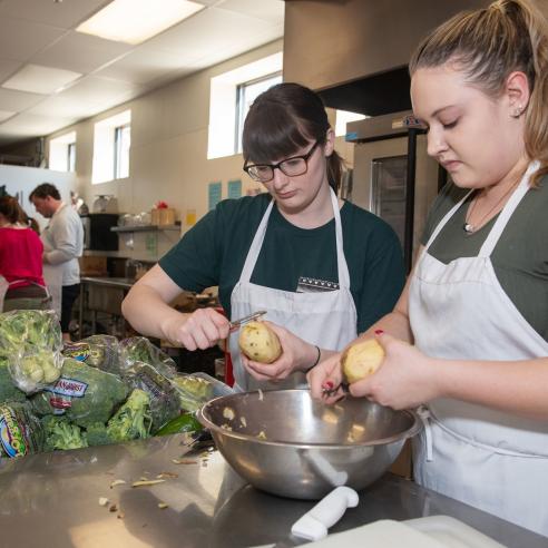 Two students at a volunteer program preparing food, peeling potatoes in a kitchen, surrounded by vegetables and other group cooking