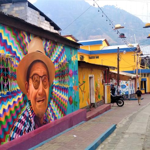 A colorful street scene in Guatemala featuring a large, vibrant mural of a smiling elderly man wearing a traditional hat, with geometric patterns and hanging hats decorating the surroundings.