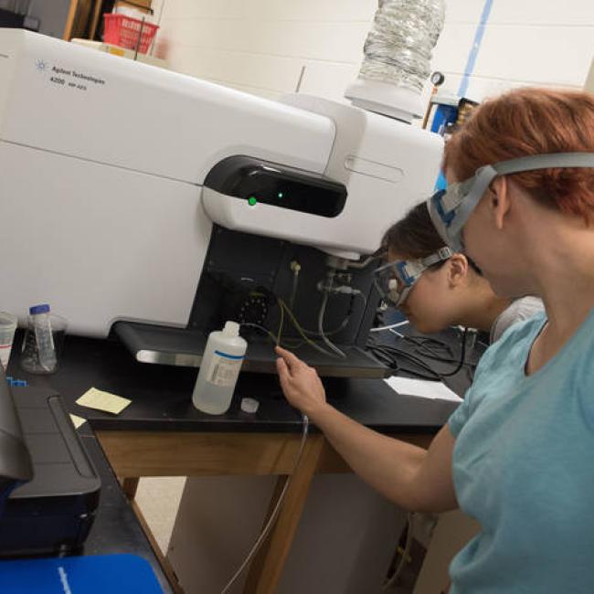 Two students wearing googles use a piece of chemistry lab equipment