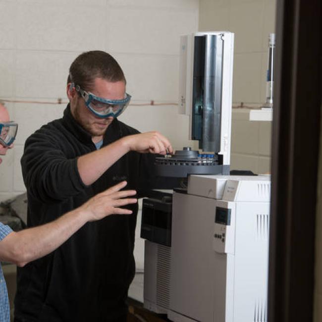 A student and a faculty member work together with chemistry equipment wearing goggles