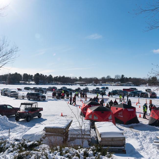 An ice fishing event on a frozen lake with participants gathered around tents and parked vehicles, enjoying winter activities in a lively, communal setting