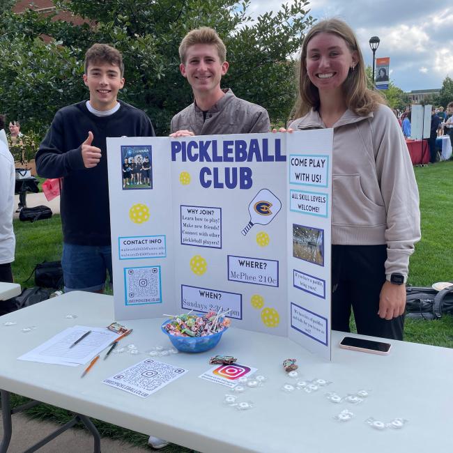 Three students stand with a Pickleball Club poster at an organization fair.