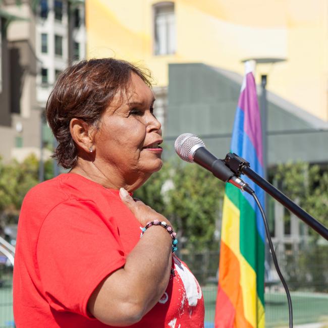 A woman wearing a red shirt, with her hand on her heart, stands aside a rainbow flag.