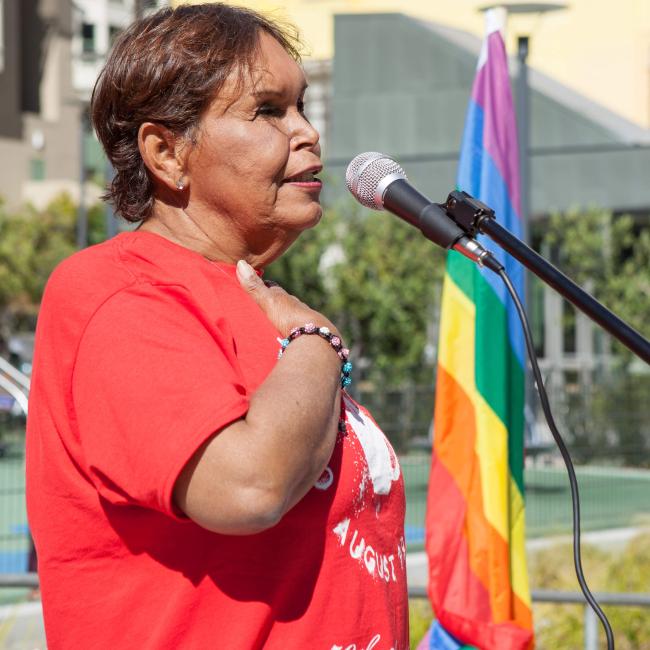 A woman wearing a red shirt, with her hand on her heart, stands aside a rainbow flag.