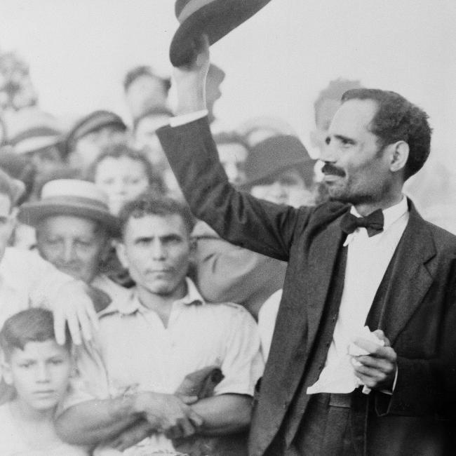 Black and white photograph of a man raising his hat to a crowd of onlookers.