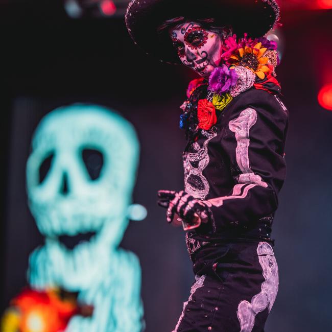 A performer on stage, dressed as el Día de los Muertos, gestures to the crowd.