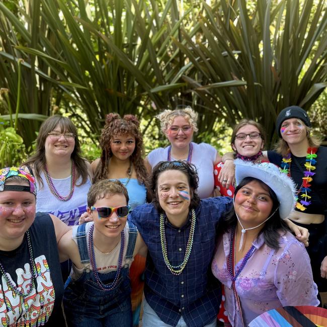 A group of students festooned in rainbow glitter and body paint smile at the camera. 