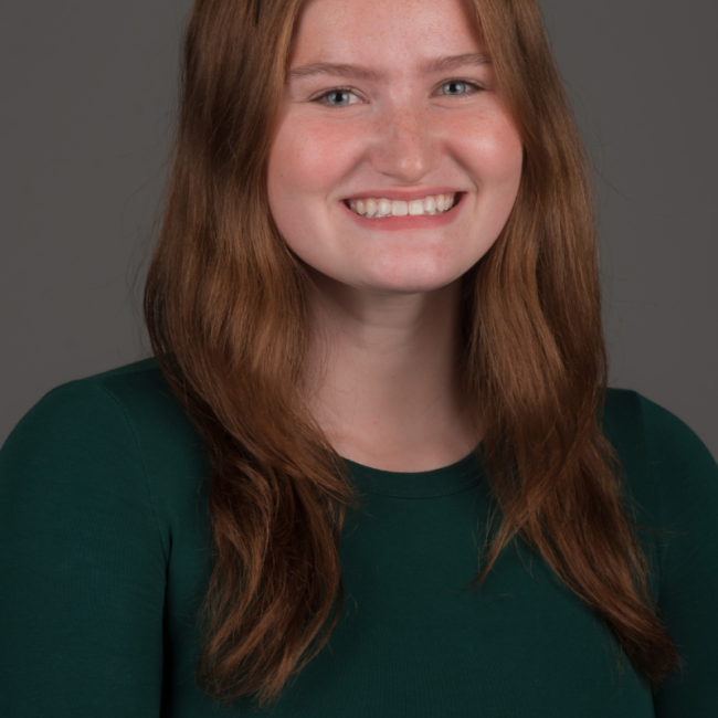 A female presenting person with shoulder length wavy red hair. She is wearing a green long sleeve top and is smiling.