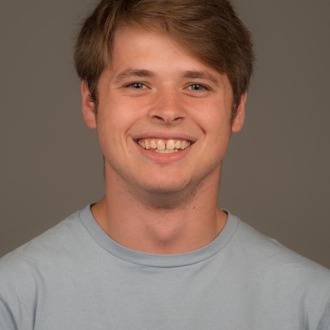 White male presenting person with short brown hair and wearing a blue T-shirt. He is smiling.