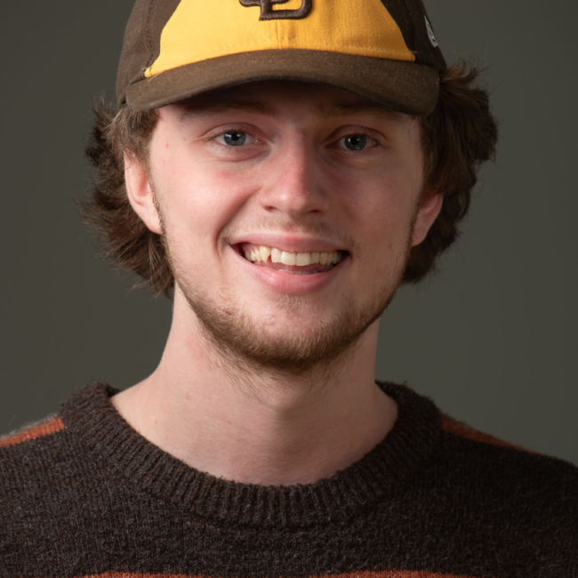 A white male presenting person wearing a brown and yellow baseball cap and striped sweater. He is smiling.