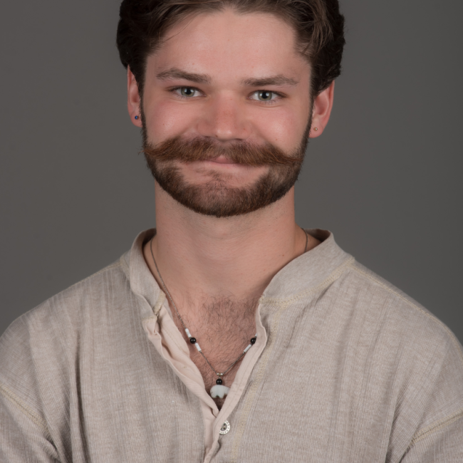 A male presenting person with short brown hair and a beard and mustache. He is wearing a beige shirt and a bear necklace.