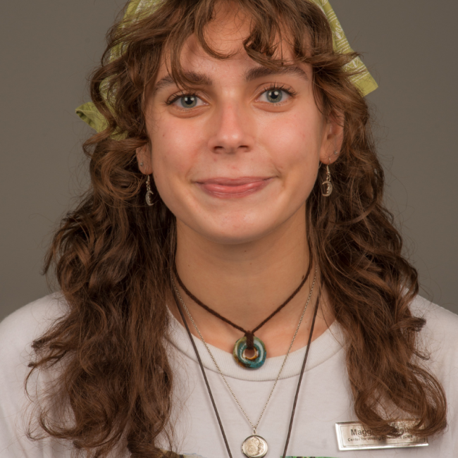 A female presenting person with long curly brown hair. She is wearing a bandanna on her head, several necklaces, and a white T-shirt. She is smiling.