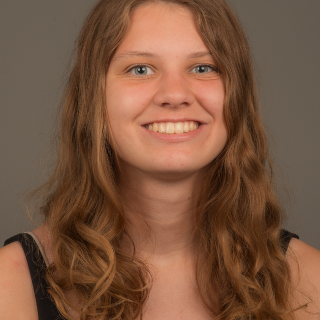 A white female presenting person with long wavy brown hair. She is wearing a black sleeveless shirt and is smiling.