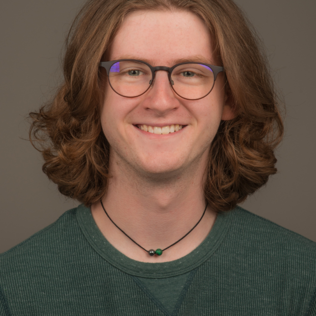 A white male presenting person with chin length curly hair. He is wearing glasses, a necklace, and green shirt. He is smiling.