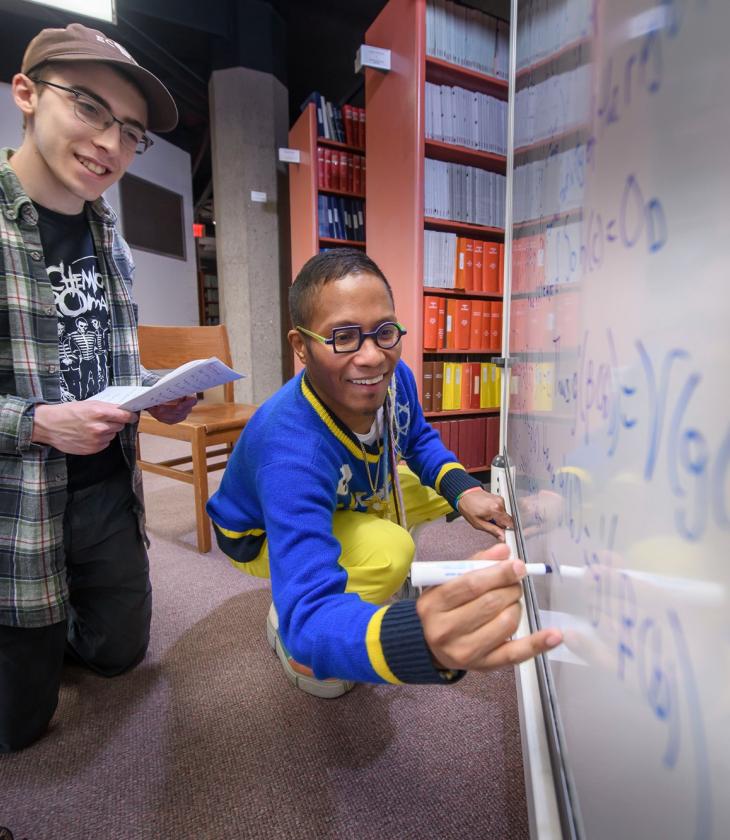 A professor and a student work out a math problem together on a whiteboard in the library.