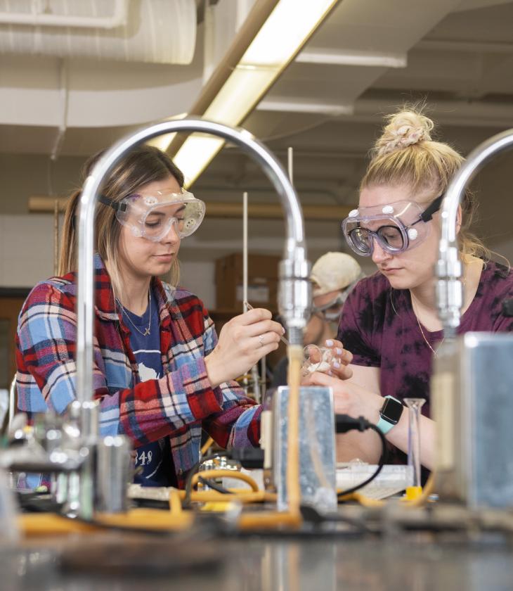 Students wearing goggles conduct an experiment in a chemistry lab.