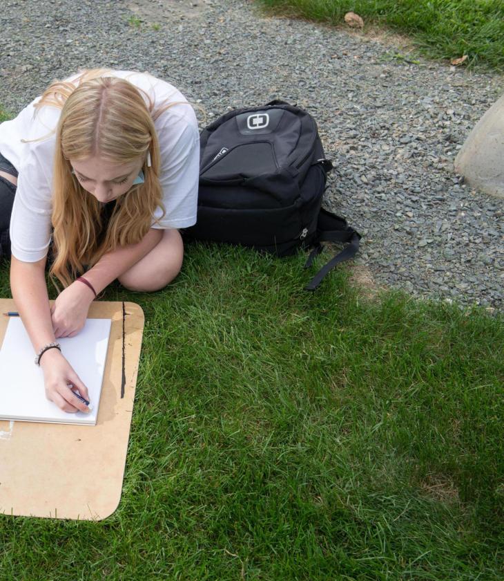 Student draws using a clipboard in the grass for an outdoor art class