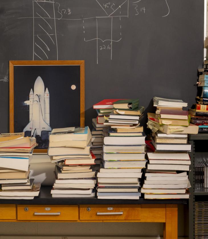 A stack of physics textbooks in front of a chalkboard and an image of a rocketship.